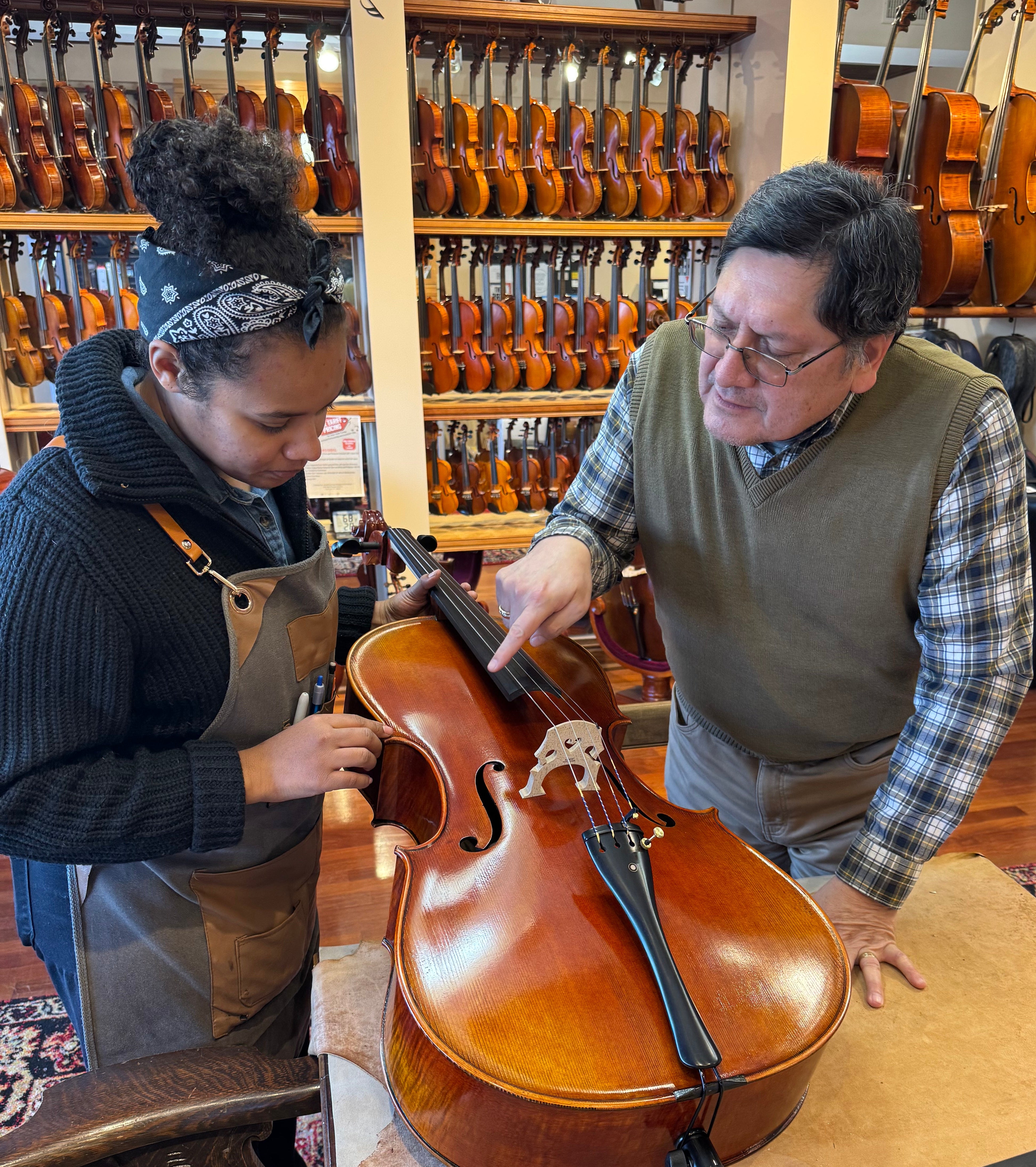 Two people examining a cello in a music store with violins on display.