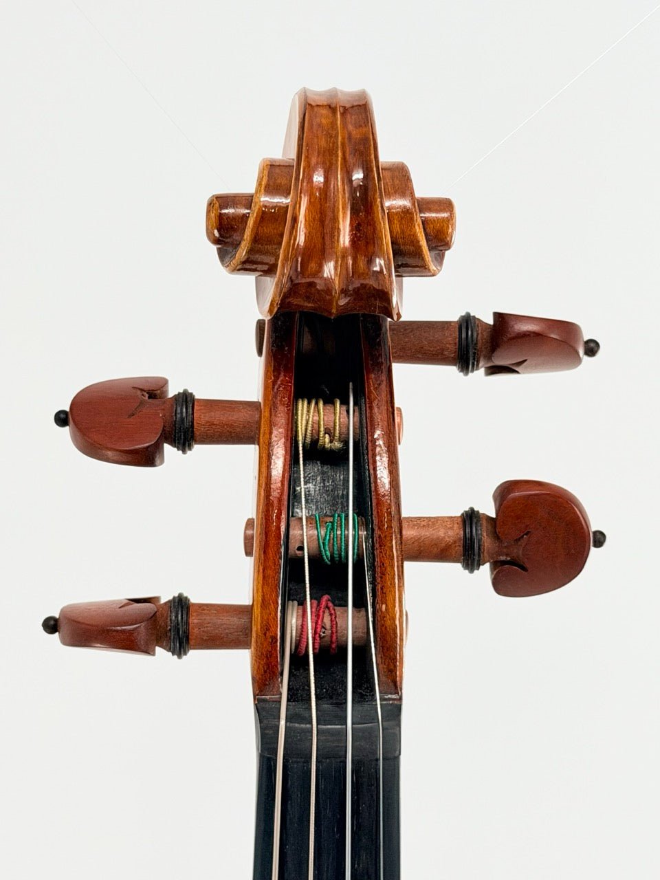 Close-up of a handcrafted violin scroll and pegbox with four wooden tuning pegs featuring colorful string windings, the polished brown headpiece shining against a white background—part of the Atelier Violin Outfit Special.