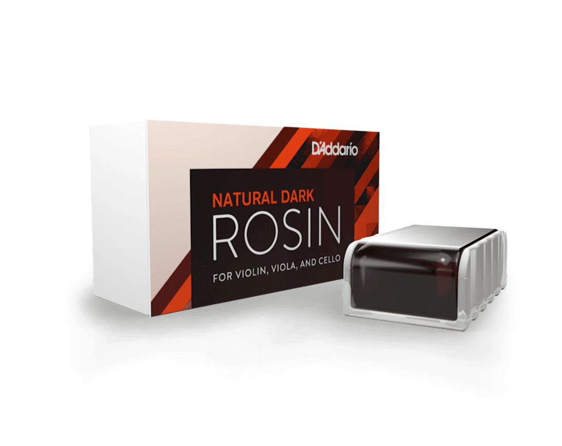 A rectangular box of D'Addario Natural Rosin Dark for violin, viola, and cello is shown next to a dark amber rosin cake in a clear holder. The packaging has bold black, orange, and white accents.