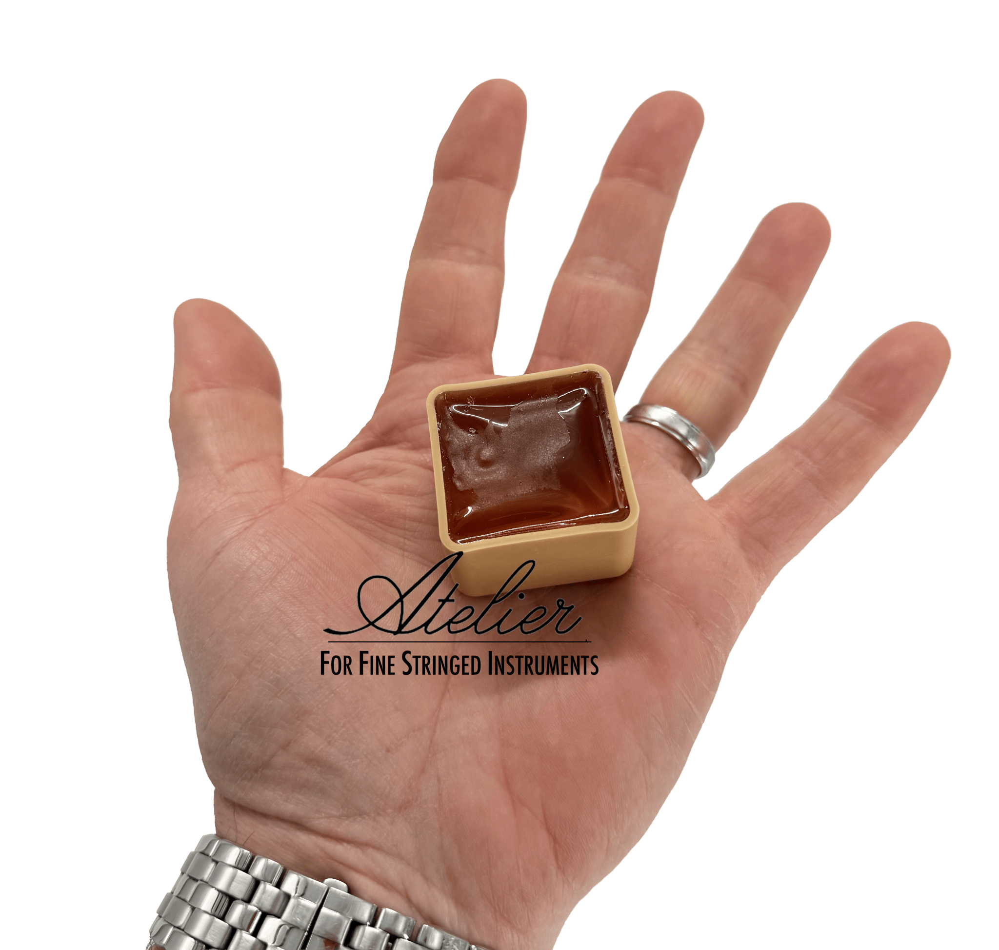 A hand holds a small square container of Eastman Bass Rosin by Leatherwood, overlaid with the text “Atelier For Fine Stringed Instruments.” The hand wears a silver watch and ring against a white background.