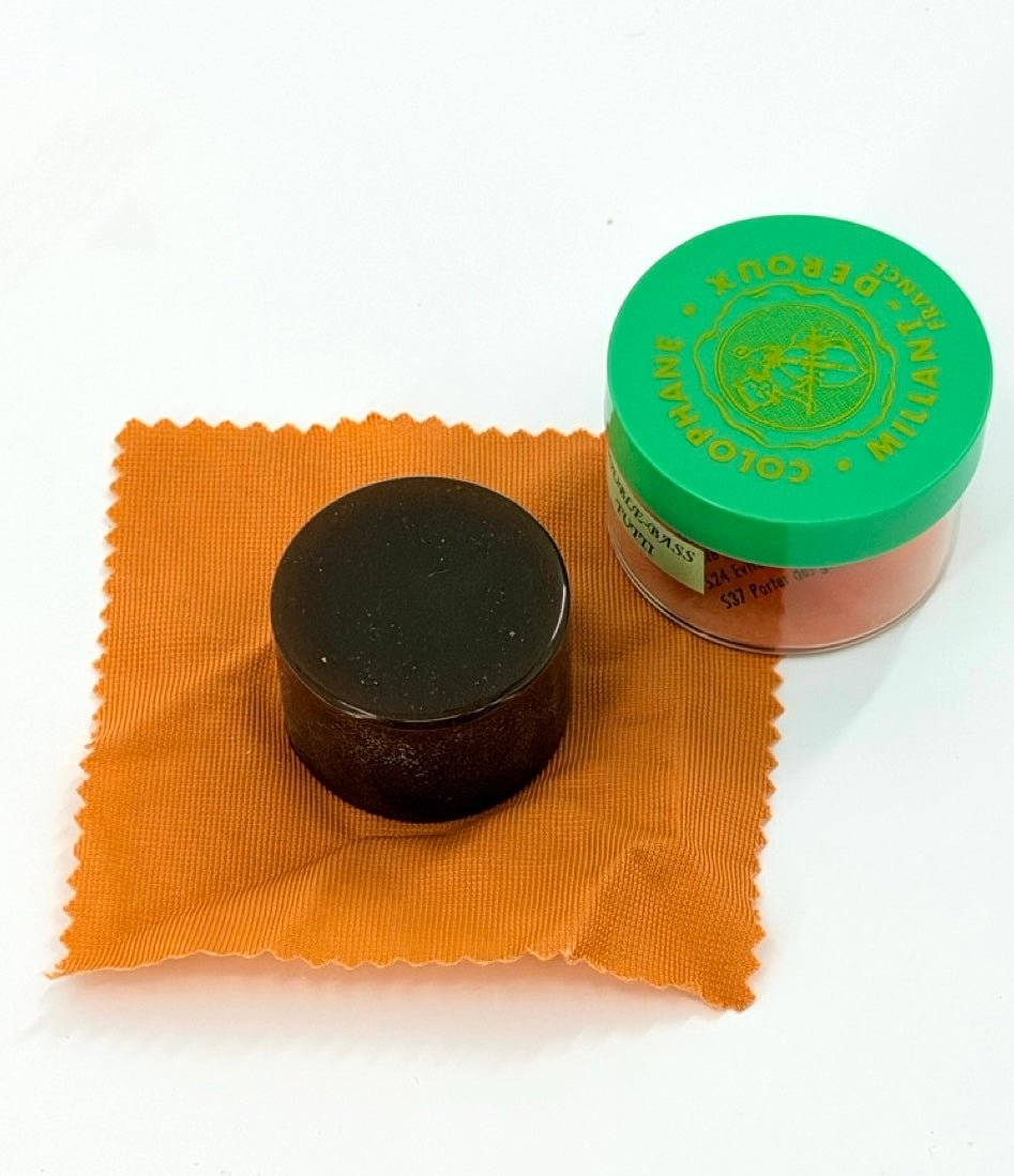 A round block of Millant-Deroux “Tutti” Double Bass Rosin sits on an orange cleaning cloth beside a round green-lidded container labeled Goldbrokat Rosin.