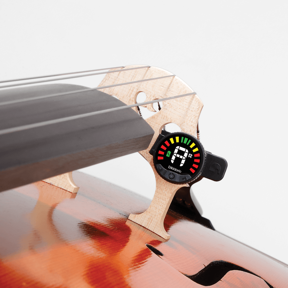 Close-up of a cello bridge with the Nexxus 360 Cello & Bass Rechargeable Tuner attached, displaying a tuning meter with red, yellow, and green indicators. The body of the cello is partially visible.