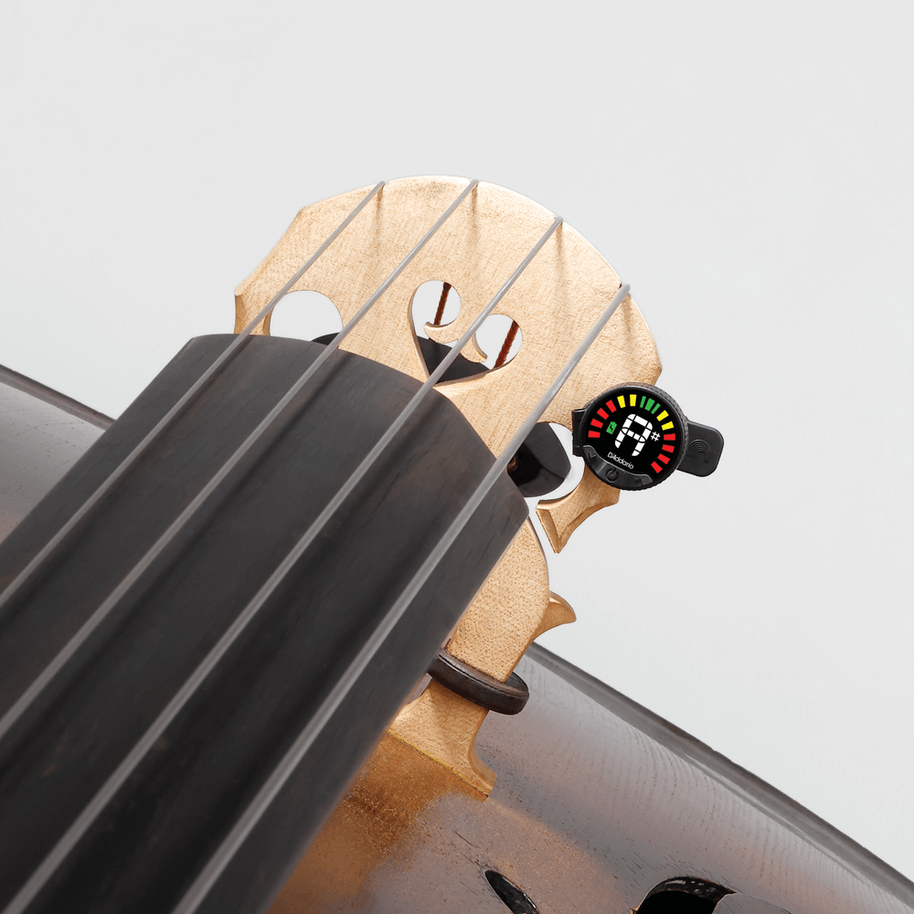 Close-up of a string instrument, likely a cello or double bass, showing the fingerboard, bridge, and a Nexxus 360 Cello & Bass Rechargeable Tuner attached near the bridge with colorful LED indicators.