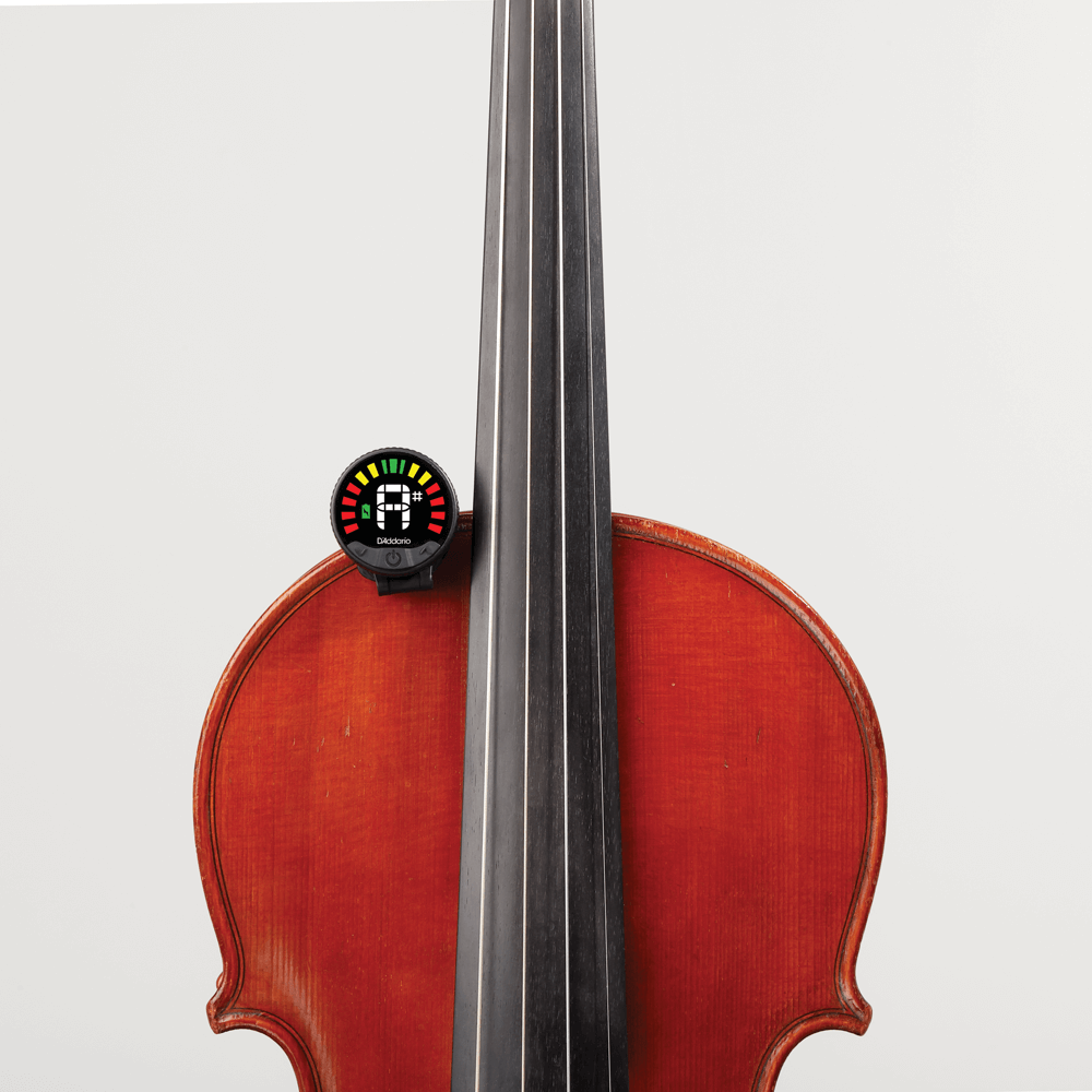 A cello with a Nexxus 360 Violin Rechargeable Tuner clipped to its side. The tuner displays a colorful meter with green, yellow, and red bars to indicate pitch accuracy. The background is plain and light gray.