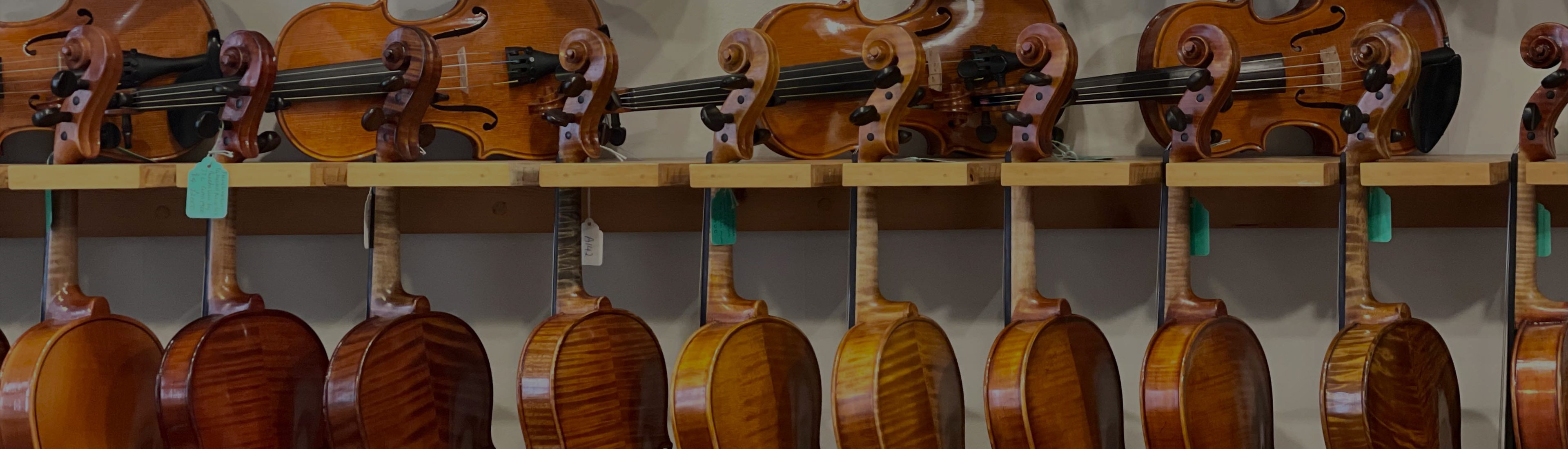 A row of violins hanging on a wooden rack, with their backs and necks visible. More violins are displayed above the rack, showing their strings and fingerboards.
