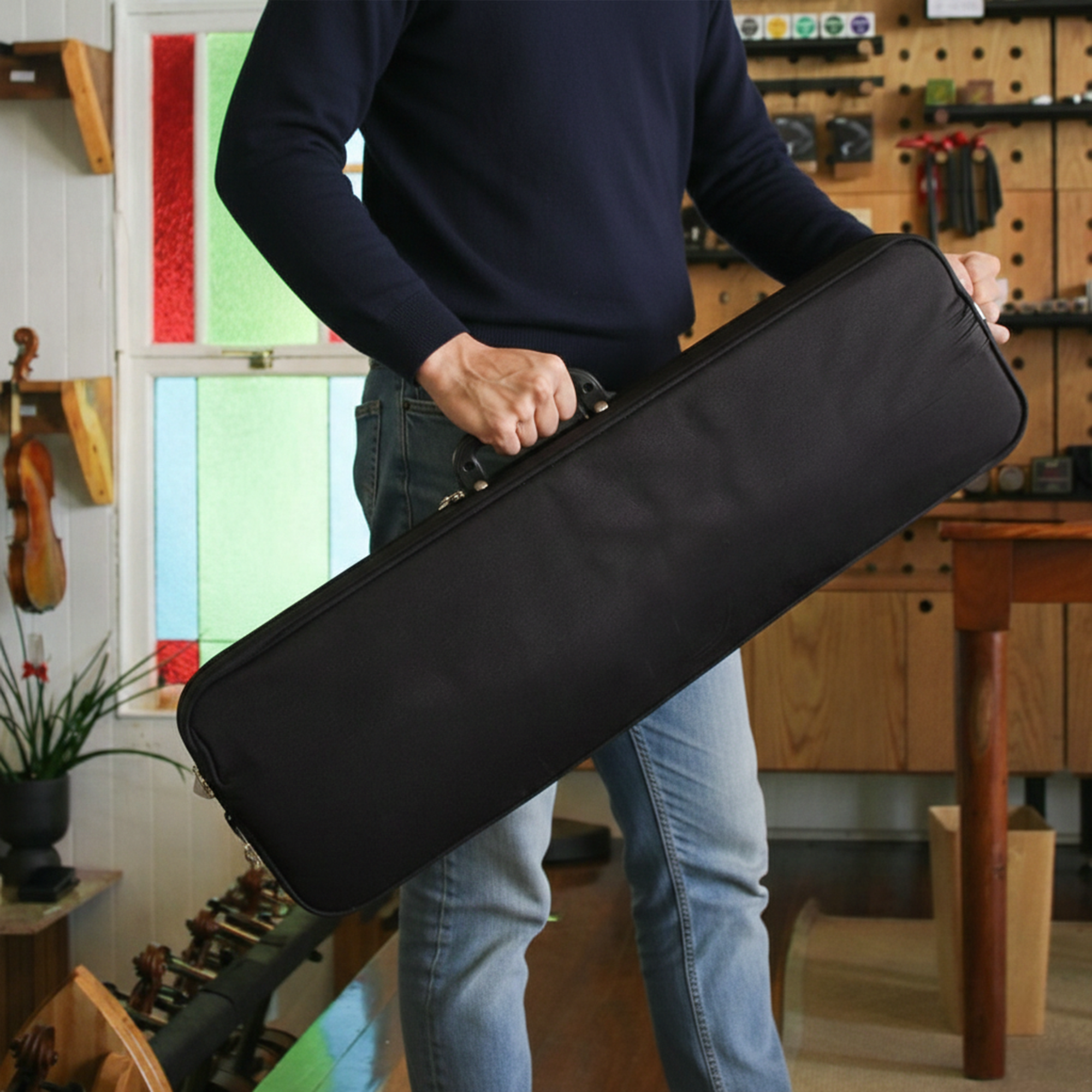 A person wearing a navy sweater and jeans holds a black rectangular instrument case in a room with wooden cabinets and string instruments on the walls.