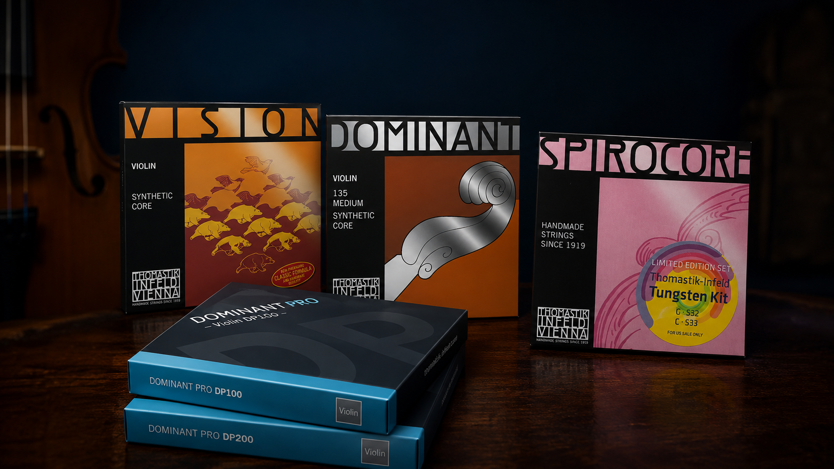 Three boxes of violin strings labeled Vision, Dominant, and Spirocore stand upright behind two flat Dominant Pro DP100 and DP200 violin string packets, all on a wooden surface.