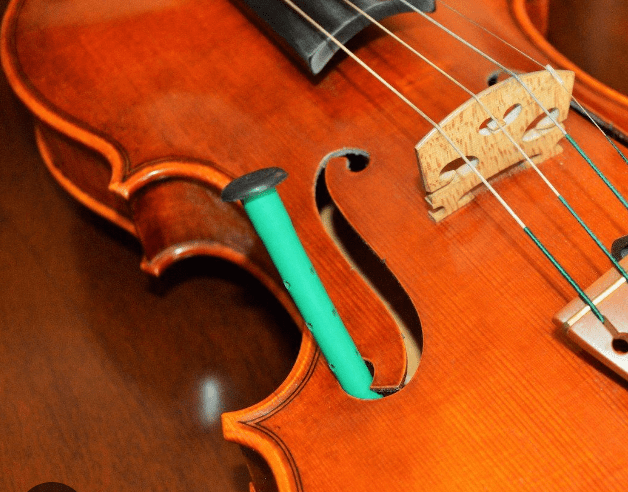 A green humidifier stick is placed inside the f-hole of a wooden violin, which is shown close up with its bridge, strings, and polished surface visible.