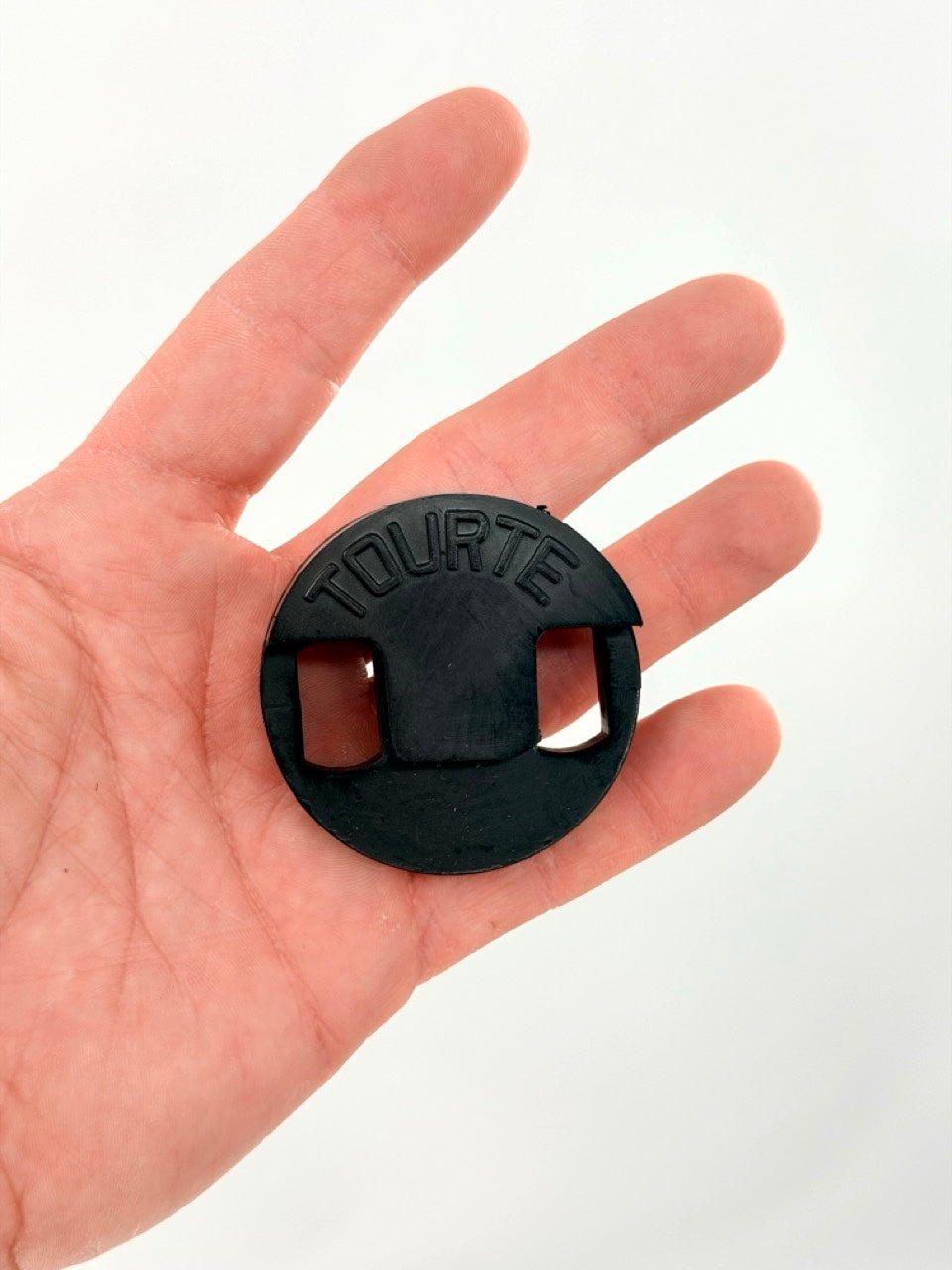 A hand holds a Tourte Bass Mute—a black rubber mute for string instruments, featuring two rectangular cutouts and "TOURTE" embossed on it. The background is plain white.