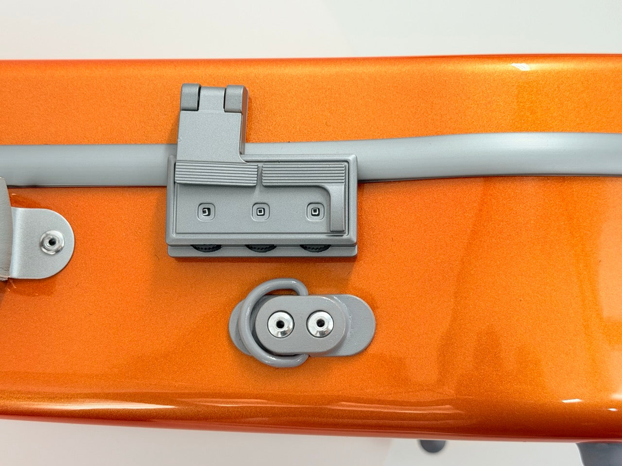 Close-up of the metallic orange surface of the Negri Eternity Carbon Fiber Shaped Violin Case – Fury Orange, showcasing its gray, futuristic latch and hinge mechanism with buttons and screws against a white background—highlighting innovative hardware characteristic of this premium violin case.