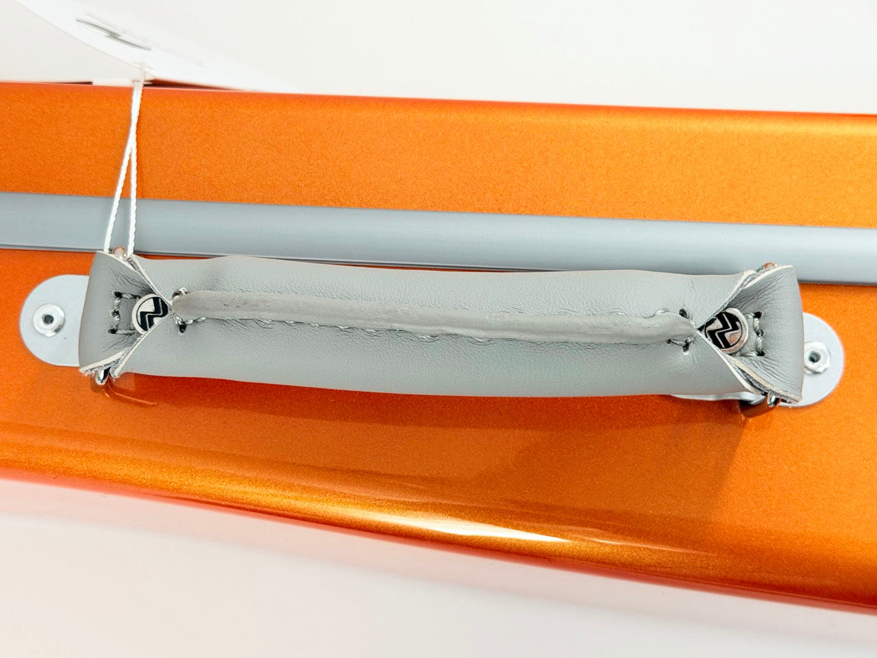 A close-up of a gray padded handle attached with string to the orange metallic surface of the Negri Eternity Carbon Fiber Shaped Violin Case in Fury Orange.