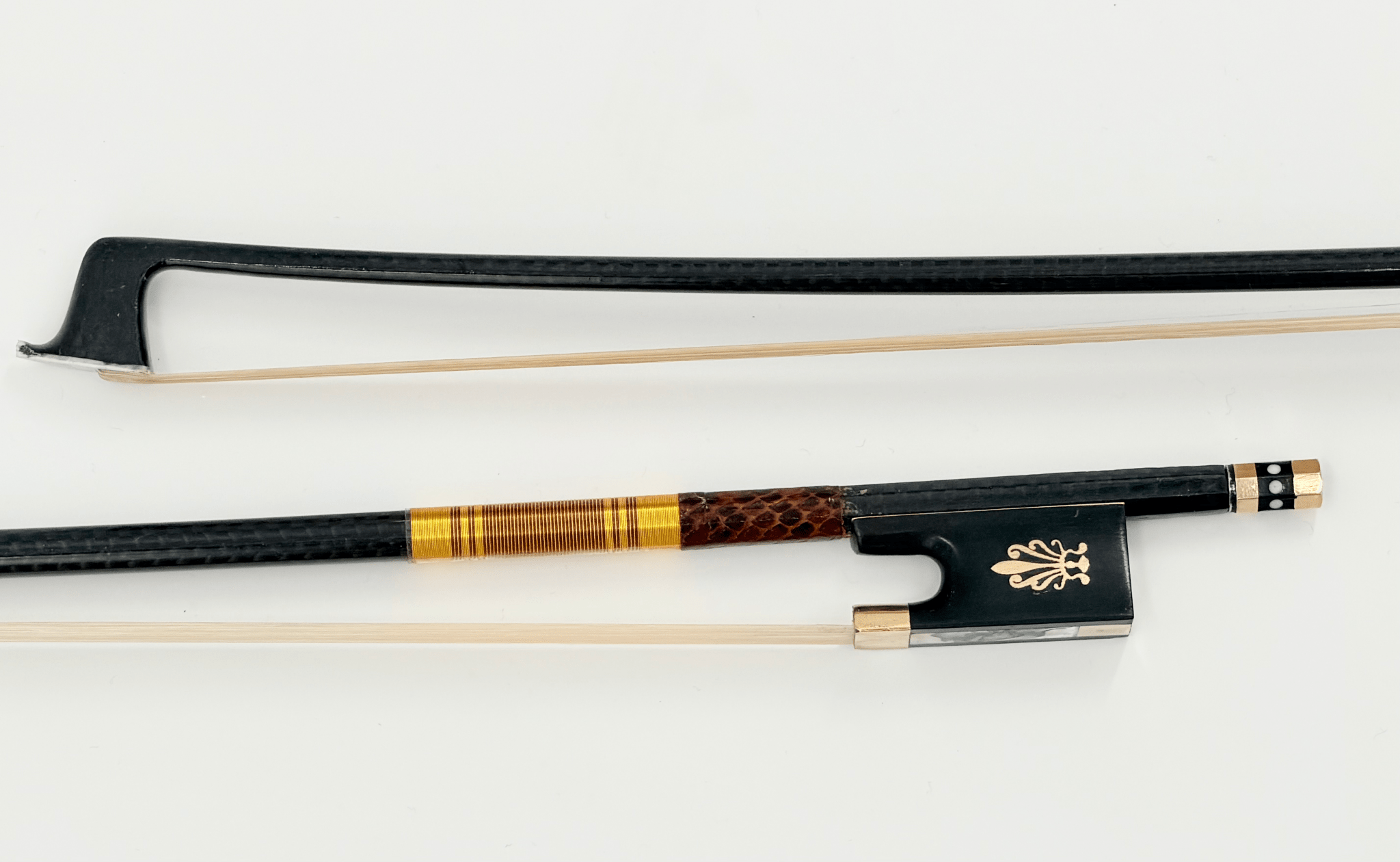 A close-up photo of the Atelier Braided Carbon Fiber Bow, showing its black stick with gold and brown detailing near the ornate gold frog design. Mongolian horsehair and the carbon fiber are clearly visible against a white background.