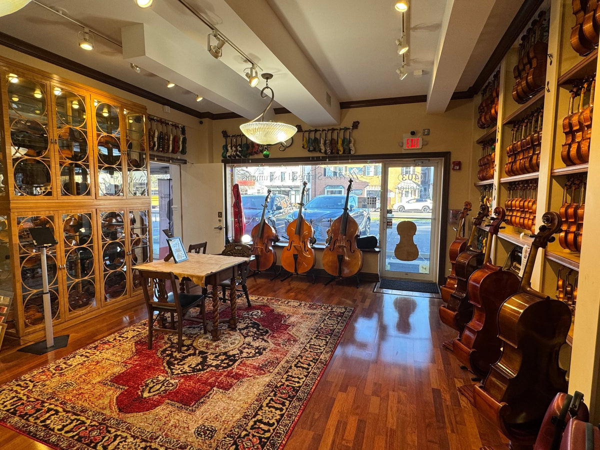 A cozy music store interior with wooden floors, violins and string instruments displayed on shelves, a table with chairs on a patterned rug, and large windows showing double basses and a street view outside.