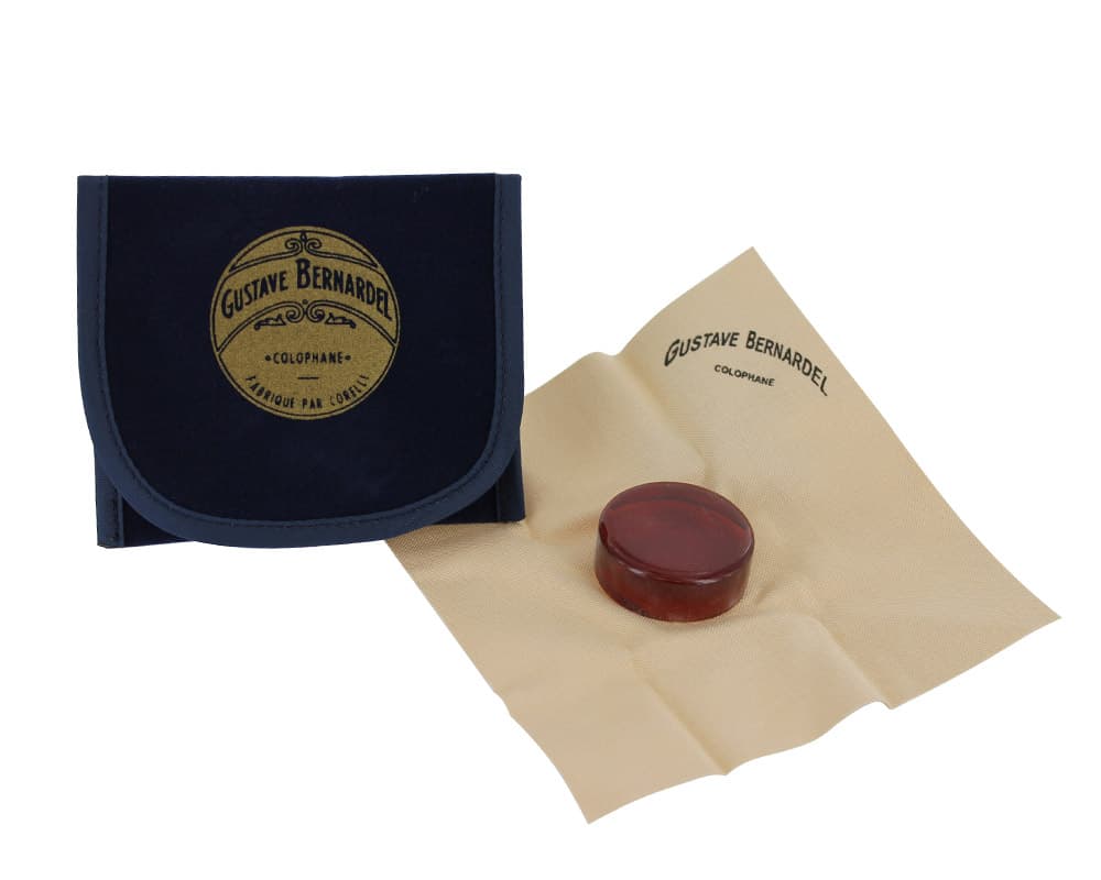 A round block of Bernardel Rosin for Violin, Viola, or Cello on a beige cloth lies next to a closed navy case labeled Gustave Bernardel Colophane.