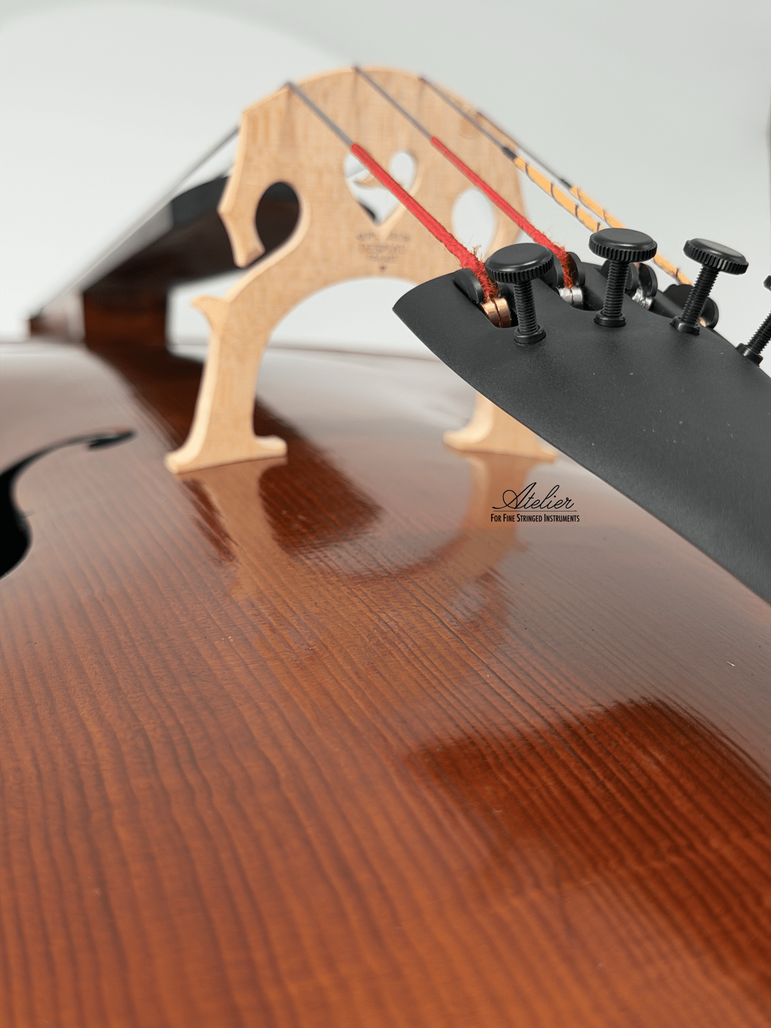 Close-up of a Juzek 303 Cello highlighting its wooden bridge, strings, and tuning pegs on a polished brown body. The Atelier brand and "Fine Tuned Stringed Instruments" are visible on this step-up cello’s surface.
