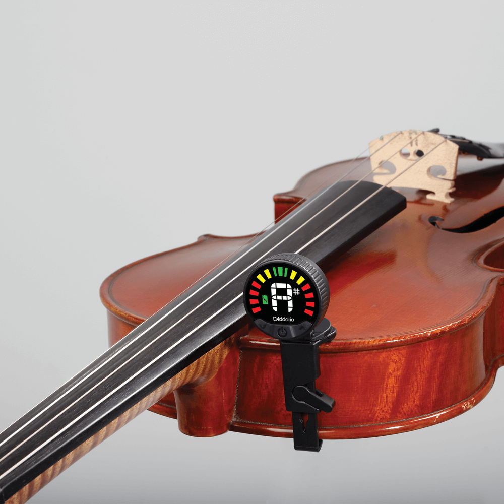 A violin with a Nexxus 360 Violin Rechargeable Tuner attached to its fingerboard, displaying colored indicator lights. The plain light gray background highlights this essential tuner for orchestral stringed instruments.