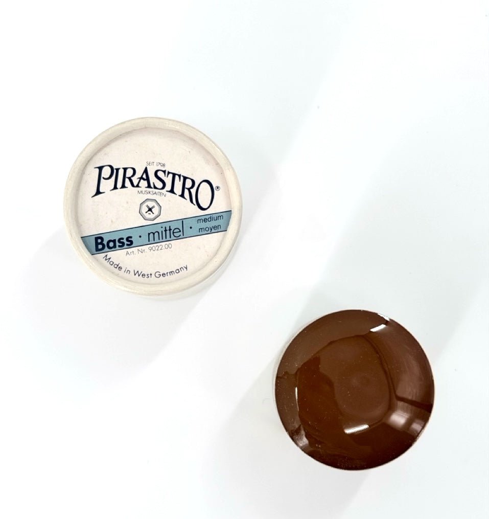 A container of Pirastro Bass Rosin – Medium sits open on a white surface, revealing the brown rosin inside. The lid displays "Pirastro Bass mittel medium moyen" and "Made in West Germany.
