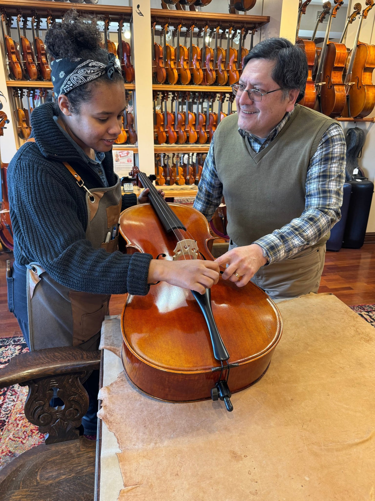 Two people in a workshop examine and work on a cello together. One wears an apron, adjusting the instrument, while the other points and smiles. Shelves filled with violins line the background wall.