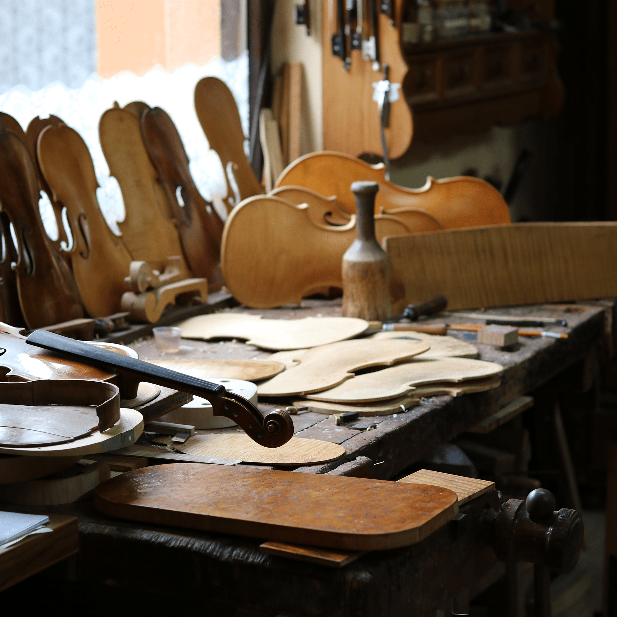 A luthier’s workshop with violin parts, wood pieces, and tools around finished and unfinished violins. Sunlight streams through a window—perfect for your Private Buying Consultation.